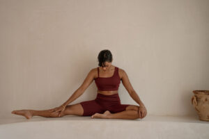 En kvinne strekker ut eller gjør yoga i et nøytralt studio. Hun har på seg et burgunder yogaantrekk og ser avslappet ut.